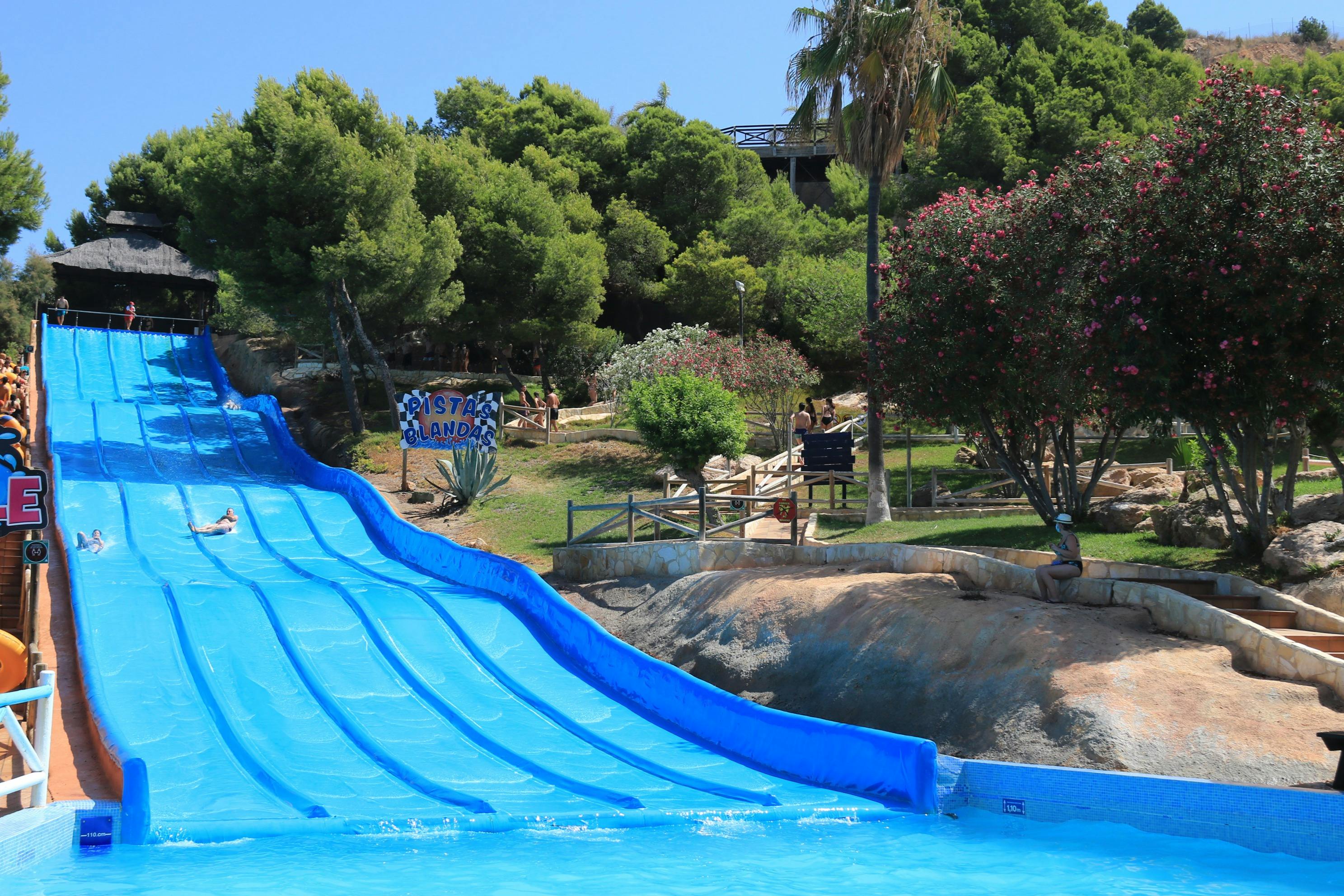 Uno scivolo acquatico blu in un parco con alberi, cespugli e, sullo sfondo, un cartello con la scritta "Aqualandia".