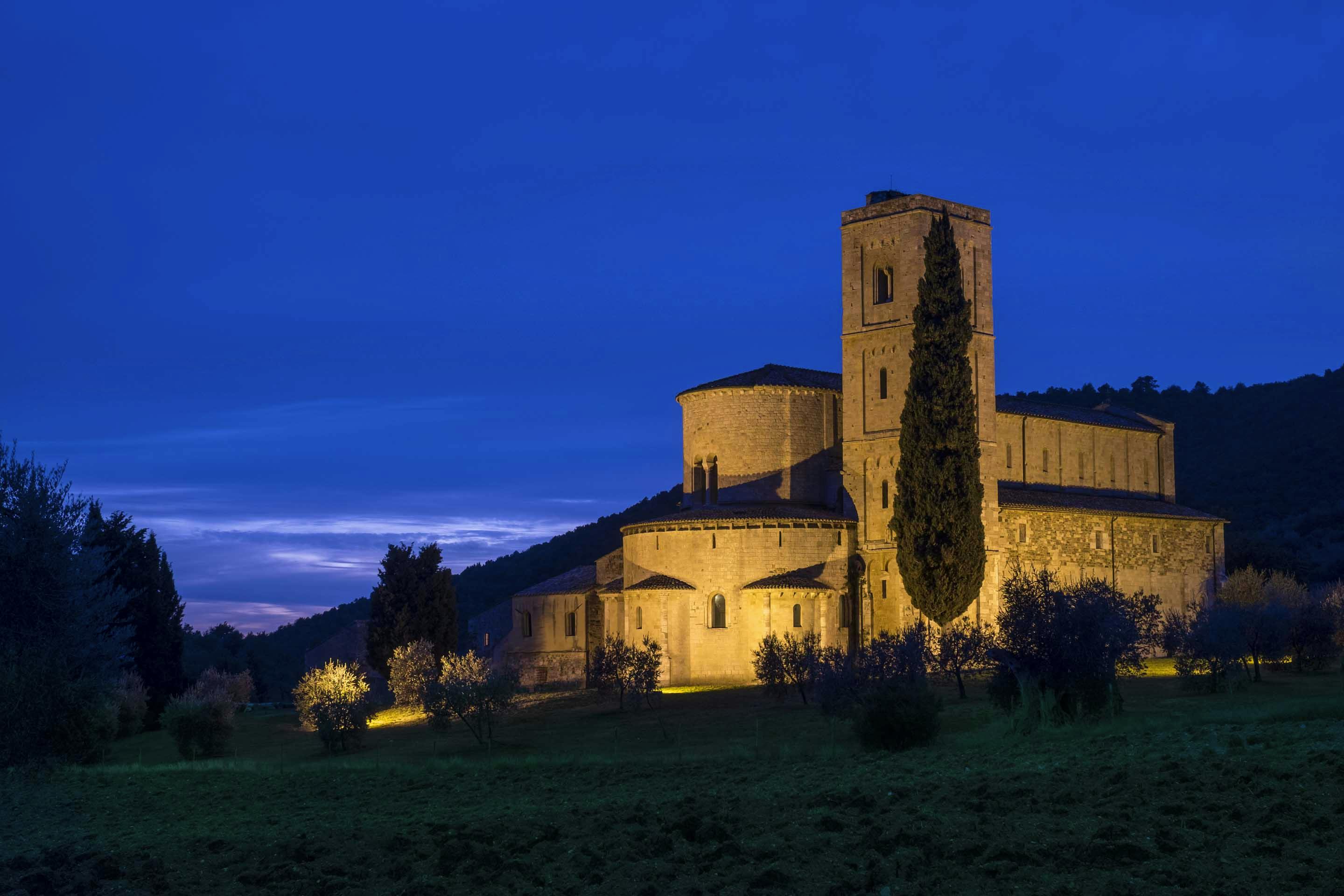 Stone church with illuminated exterior at dusk, surrounded by trees and a grassy field, under a deep blue sky.