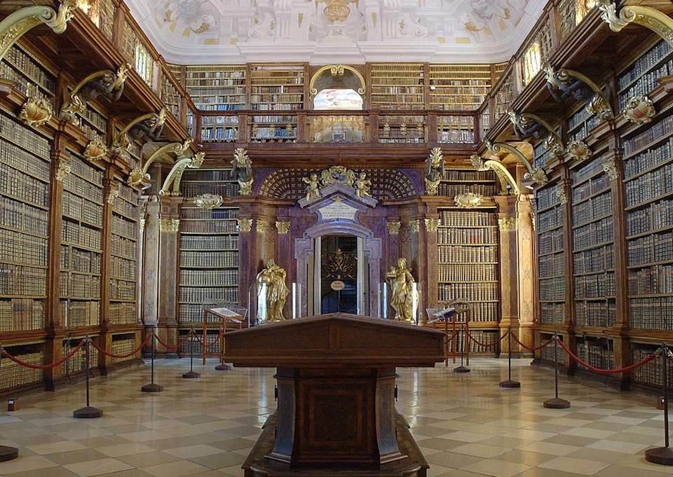 Ornate library with wooden shelves filled with books, statues, and intricate carvings. Central reading desk in the foreground.