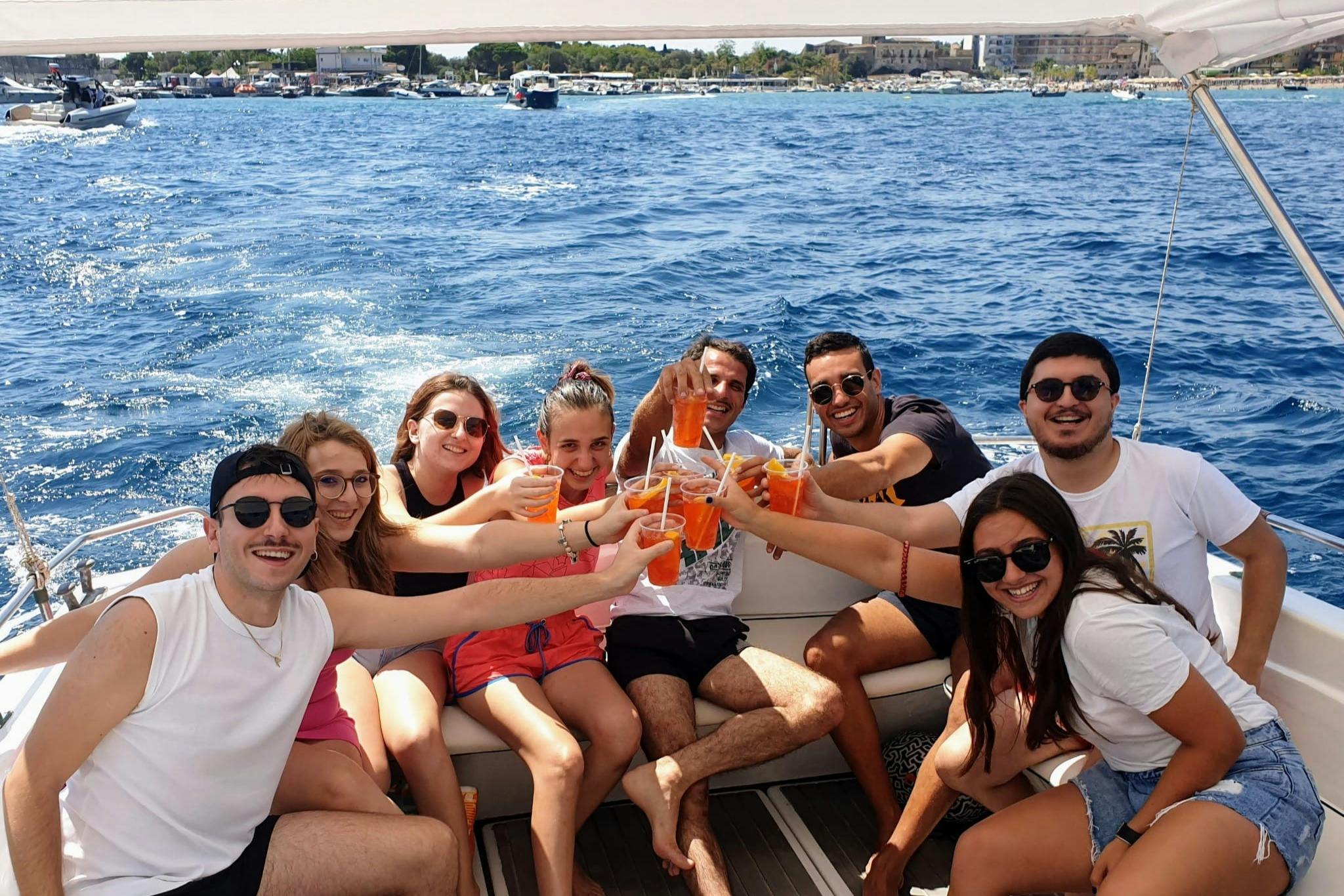 Un gruppo di persone sedute su una barca, che sorridono e alzano bicchieri con bevande arancioni, con un mare e una costa blu sullo sfondo.