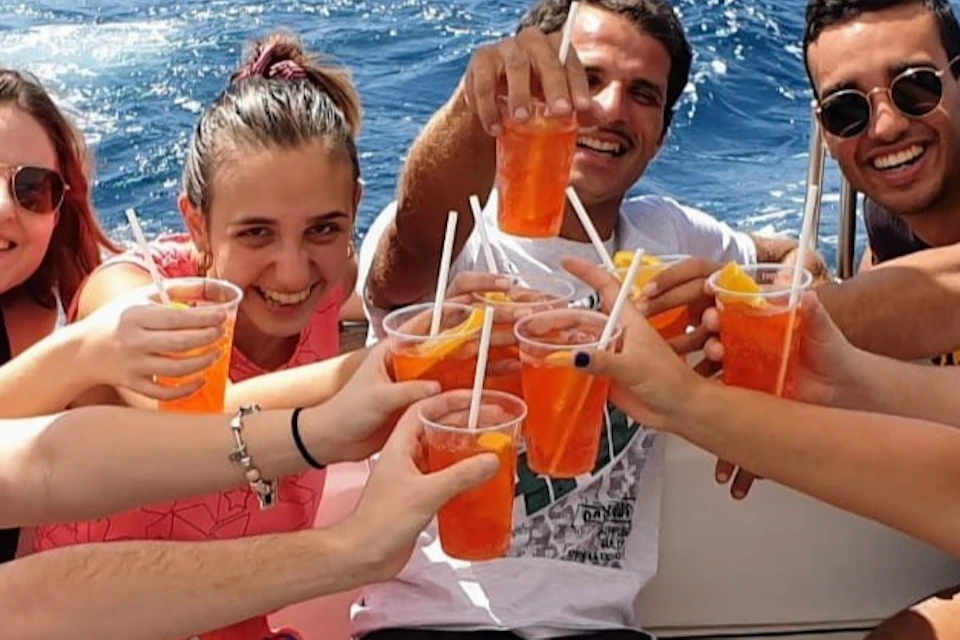 A group of people sitting on a boat, smiling and raising glasses with orange drinks, with a blue sea and coast in the background.