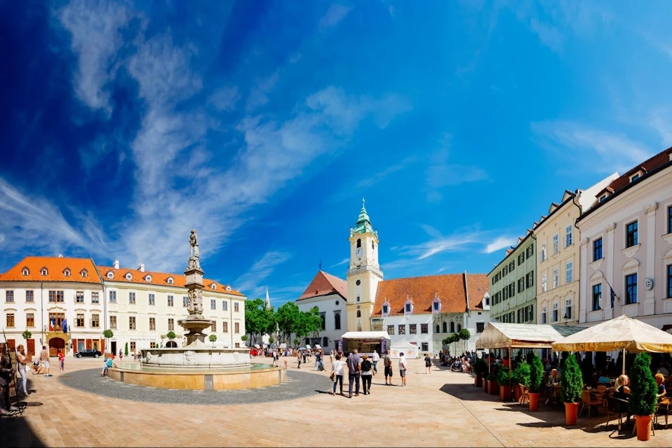 A bustling town square with a central fountain, historic buildings, and people walking under a clear blue sky.