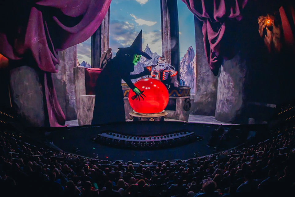 A stage performance featuring a witch with a glowing red orb and a seated figure in a fantastical setting before an audience.