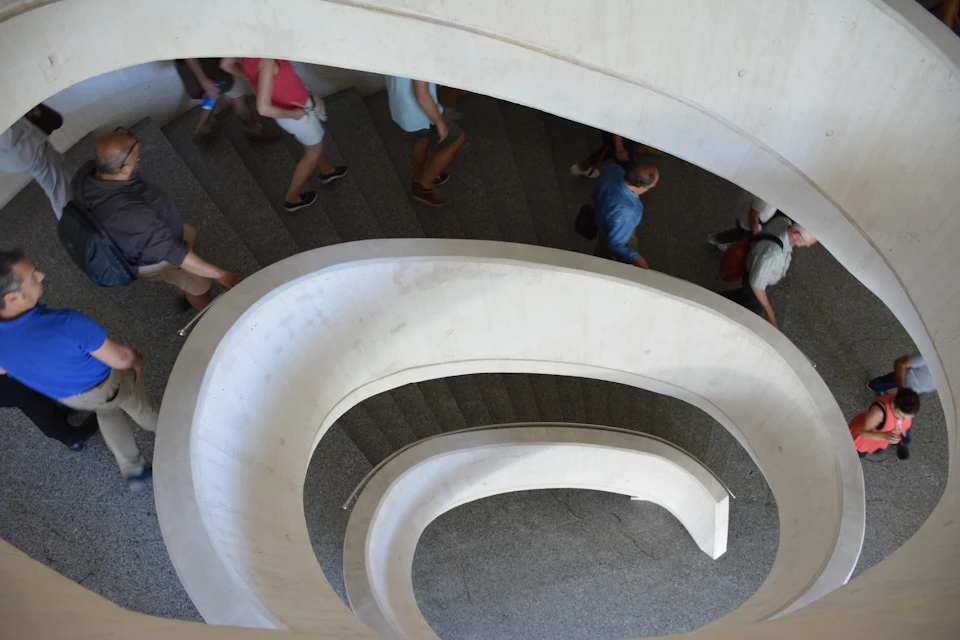 People walking up and down a curved, spiraling staircase with white railings and dark steps.