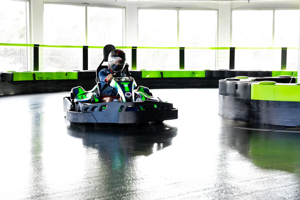 Person driving a green and black go-kart on an indoor track with reflective flooring and large windows in the background.