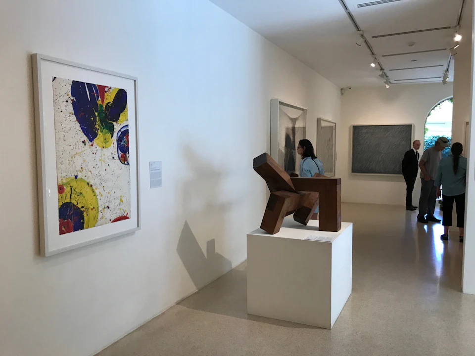 A modern art gallery with abstract paintings on the walls and a wooden sculpture on a pedestal. Two people observe the artwork.