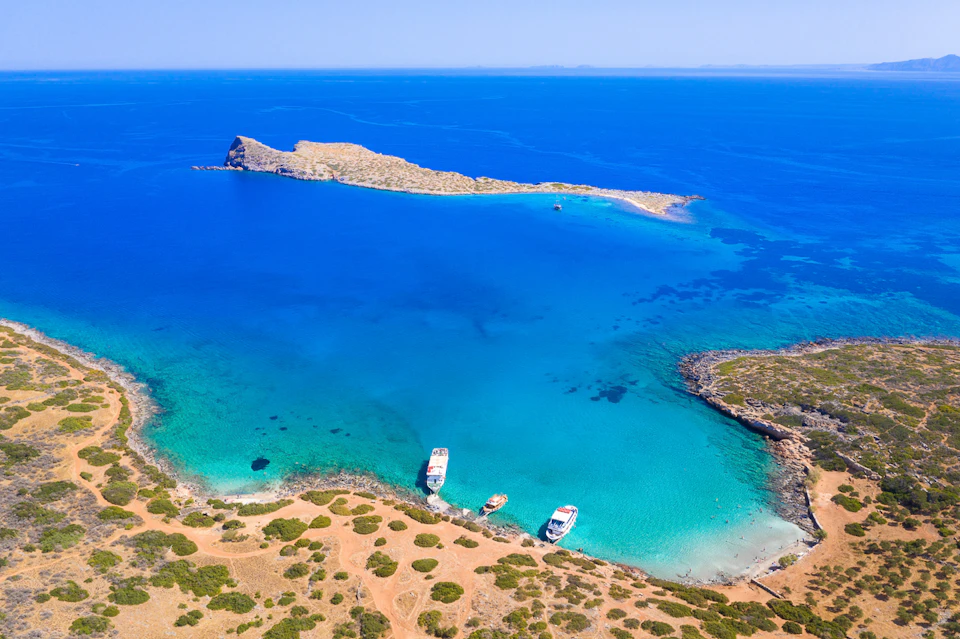 Aerial view of a vivid blue bay with three anchored boats near a rugged, sandy coastline and a small island in the background.