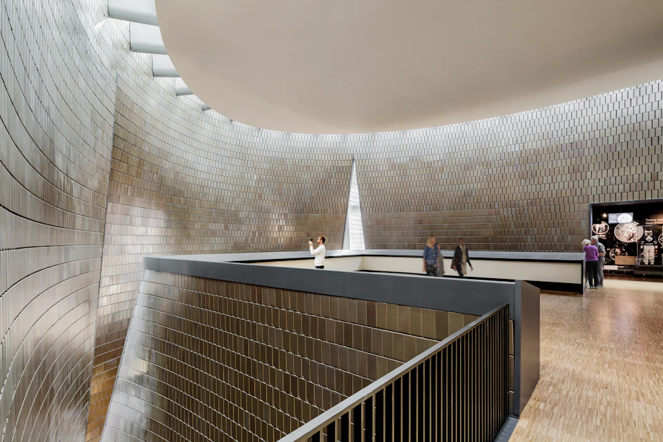 Three people walk on a balcony in a modern, spacious building with metallic tiled walls and a high, curved ceiling.