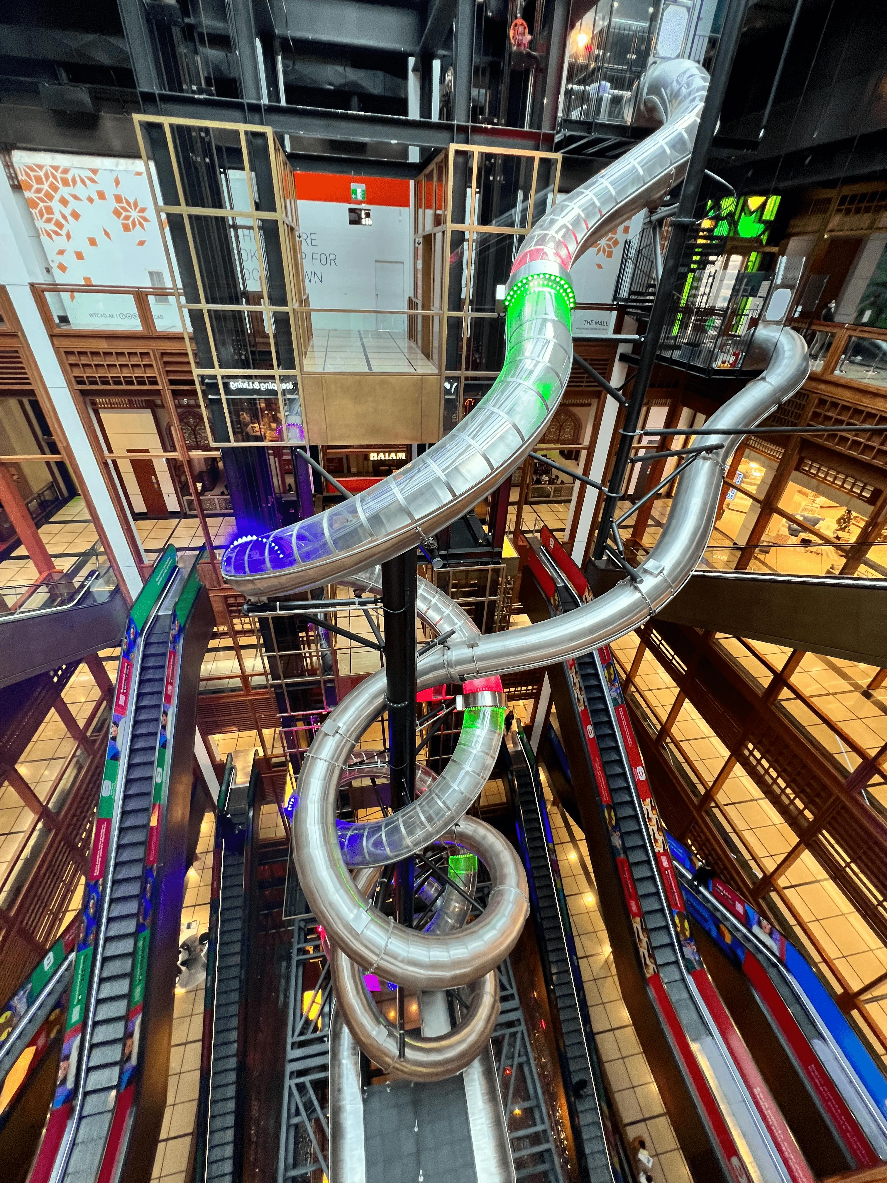Velký víceúrovňový vnitřní skluzavka s průhlednými trubicemi a barevnými světly, spirálovitě procházející atriem nákupního centra.