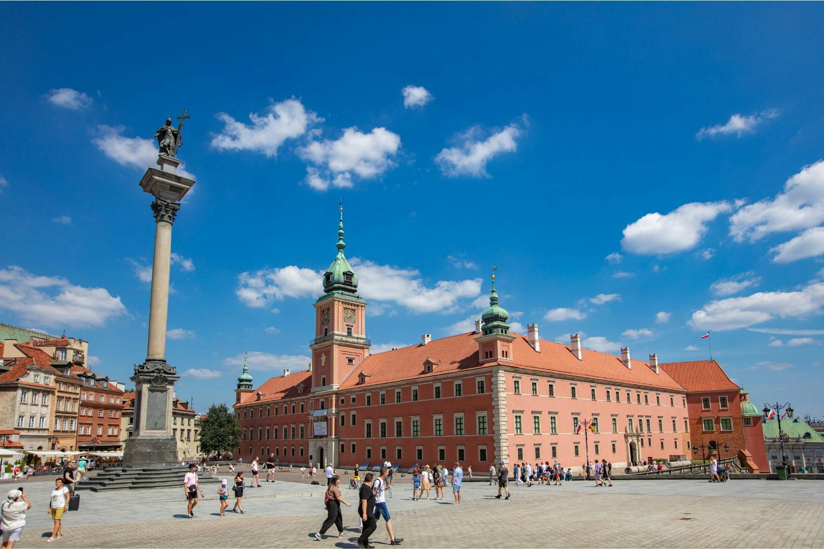 Ein historisches rotes Gebäude mit grünen Türmen unter einem blauen Himmel, eine hohe Säulenstatue und Menschen, die auf einem Platz spazieren gehen.