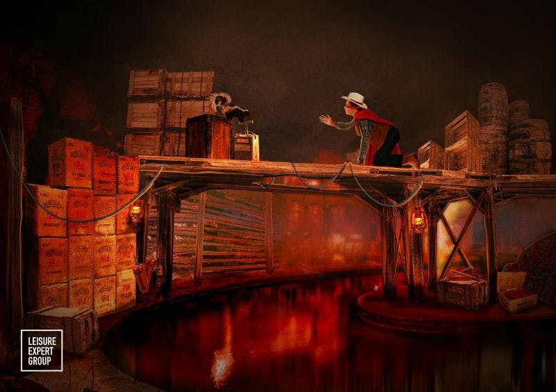 A person in a cowboy hat and red cape leans over a wooden bridge, reaching towards a raccoon next to stacked crates.
