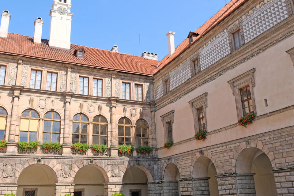 A historic courtyard surrounded by ornate stone buildings with arched windows, red-tiled roofs, and flower planters.