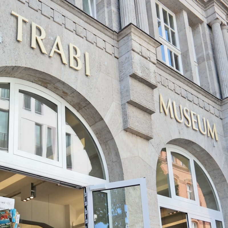 Det ydre af en bygning mærket "Trabi Museum" med store buede vinduer og en åben døråbning i bunden.