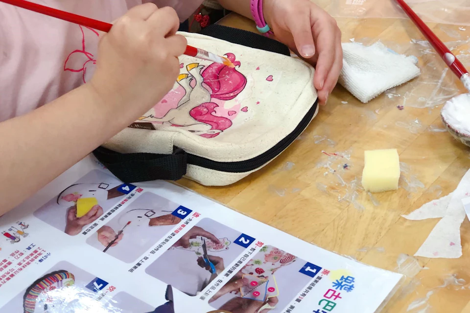 A person painting a design on a fabric pouch, with instructions for crafting visible on the table.
