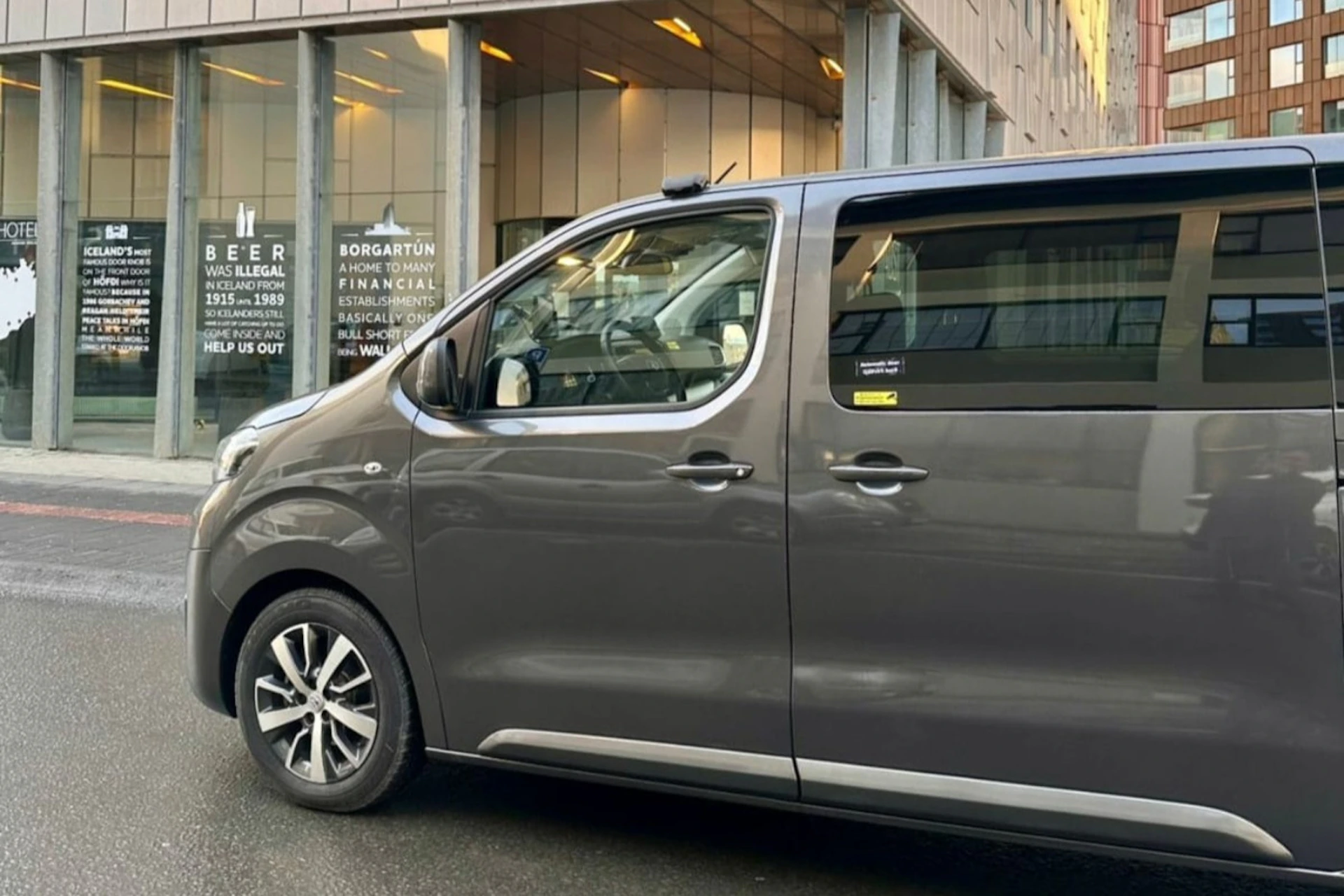 A gray minivan parked on the street in front of a glass-fronted building with signs and text visible through the windows.