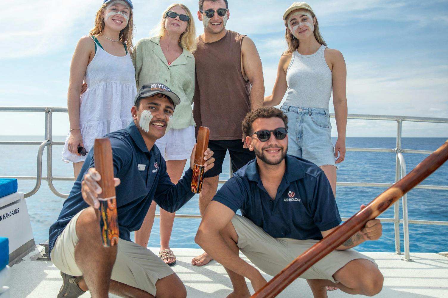 Group of five people posing on a boat, two kneeling holding wooden items, three standing, all smiling under a partly cloudy sky.