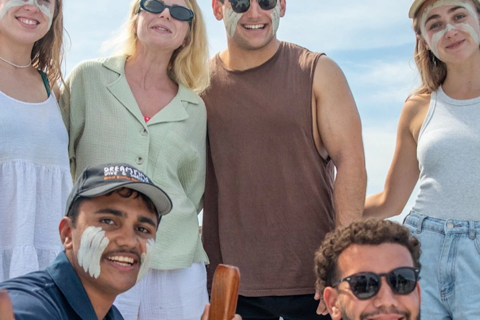 Group of five people posing on a boat, two kneeling holding wooden items, three standing, all smiling under a partly cloudy sky.