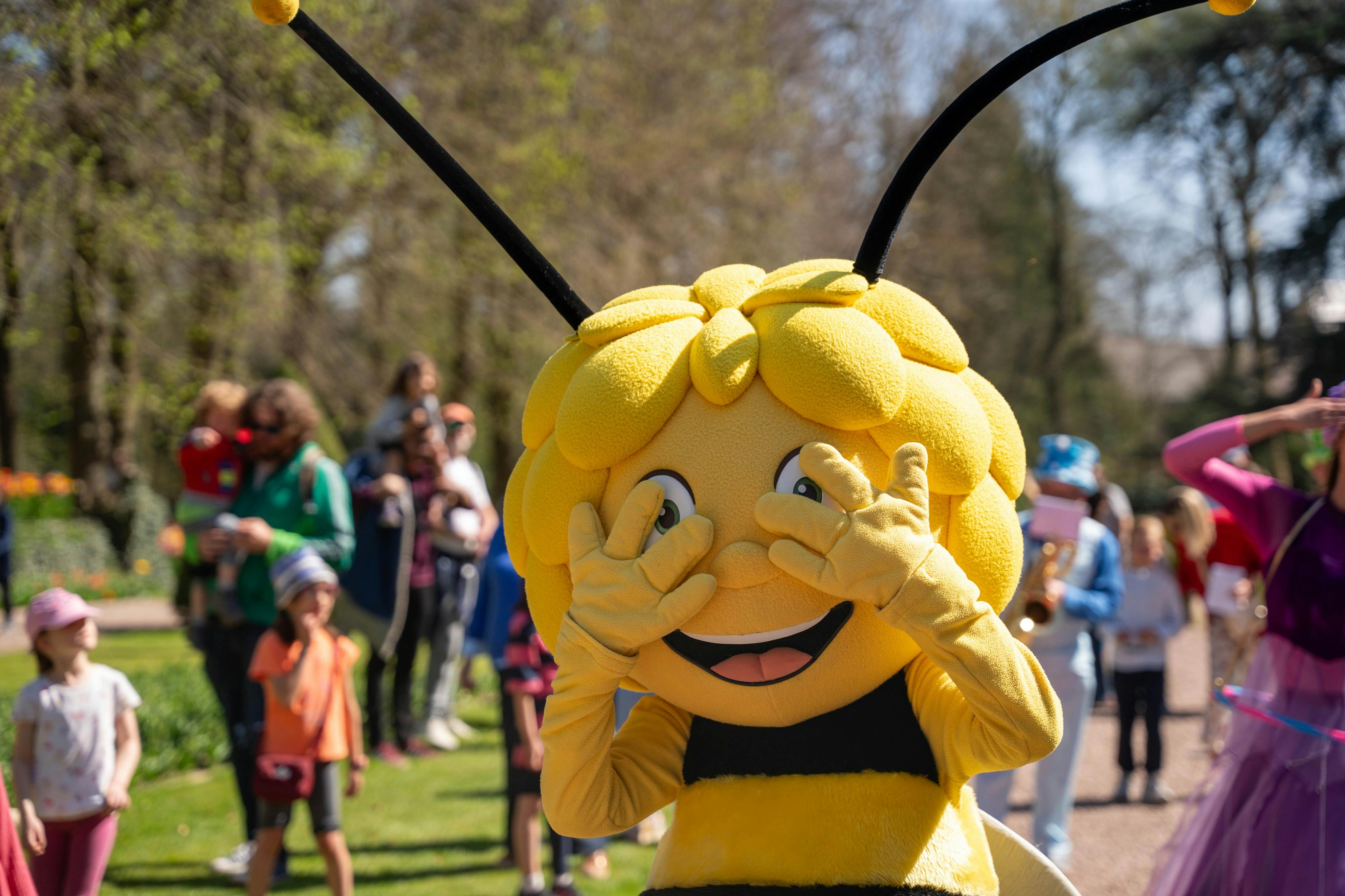 Uma pessoa vestida com um fato de abelha a posar de forma divertida ao ar livre, com pessoas e árvores ao fundo num dia de sol.