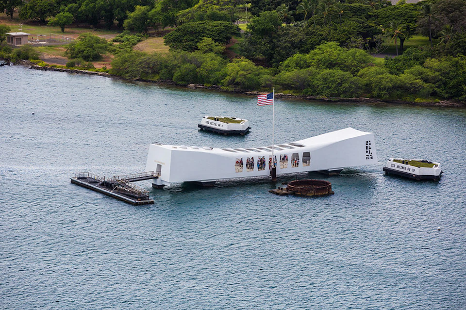 White memorial structure over water, American flag, and boat nearby. Green vegetation in the background.