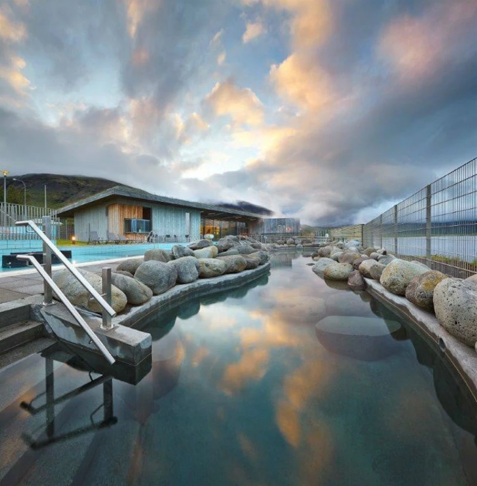 A serene hot spring with large rocks, a wooden building, and a pool ladder under a dramatic sky with clouds reflecting in the water.