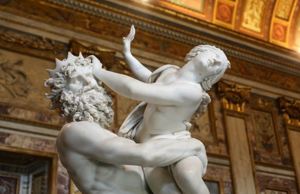 Marble statue of a bearded man holding a struggling young woman in a room with ornate walls and ceiling.