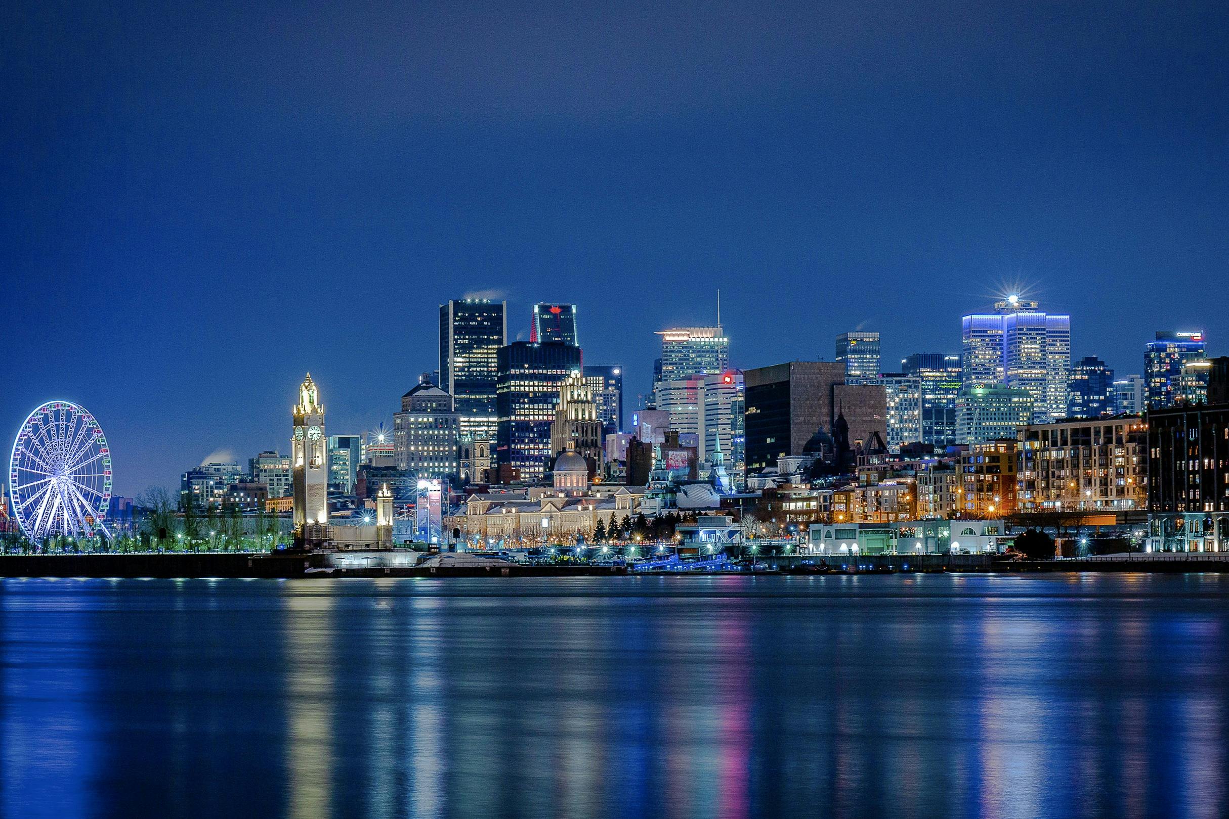 City skyline at night with illuminated buildings reflecting on a calm body of water.