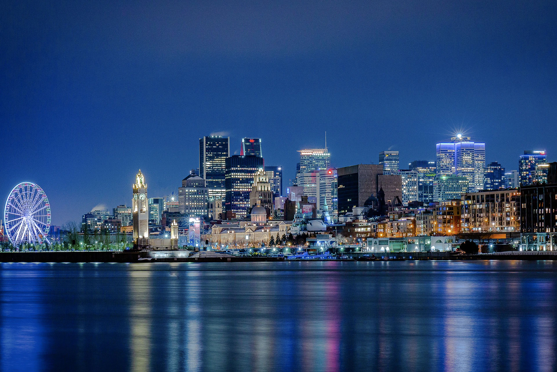 City skyline at night with illuminated buildings reflecting on a calm body of water.