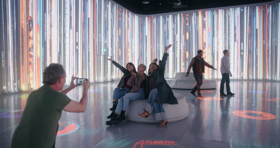 Iemand maakt een foto van drie mensen die poseren tegen een achtergrond van kleurrijke, verticale digitale beeldschermen in een overdekte omgeving.