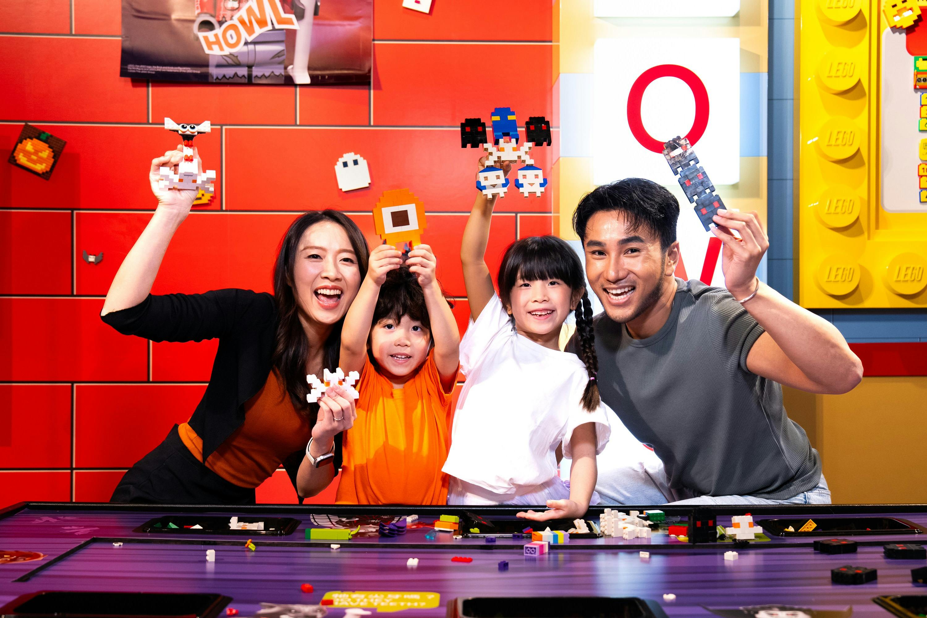 A family of four smiling and holding small LEGO figures at a building station with colorful LEGO pieces on the table.