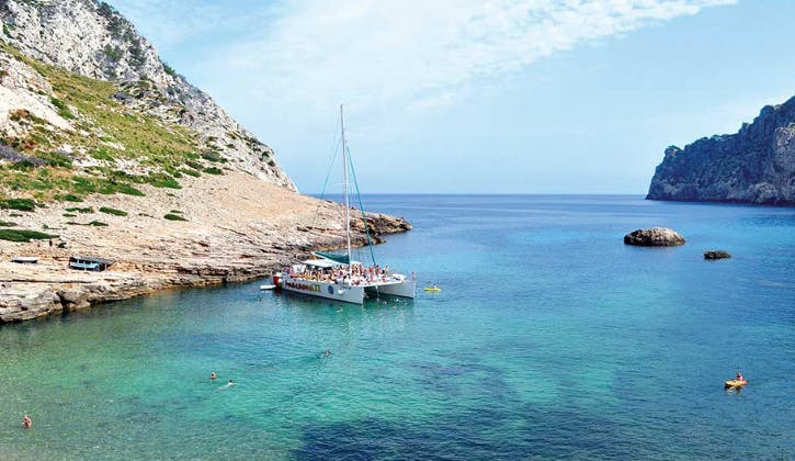 Un catamaran avec des personnes à bord est ancré près d'une côte rocheuse avec une eau bleue claire. Des nageurs sont visibles à proximité.