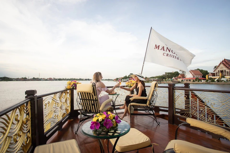 Two women seated on a boat, clinking glasses near a table with flowers. A "Manohra Cruises" flag is visible.