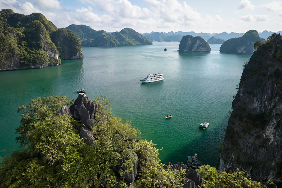 Aerial view of a calm bay with several boats surrounded by lush, rocky islands under a bright blue sky with clouds.