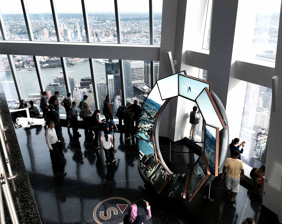 People viewing a cityscape from an observation deck with large windows and a circular display in the foreground.