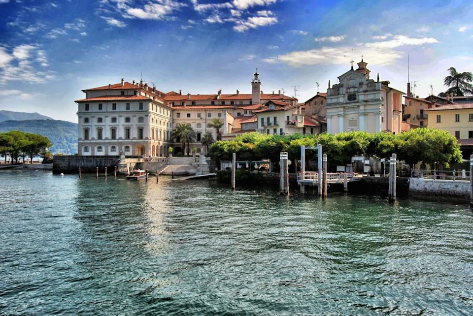 Historic buildings with red roofs and a white church by a waterfront with several docks, under a partly cloudy sky.