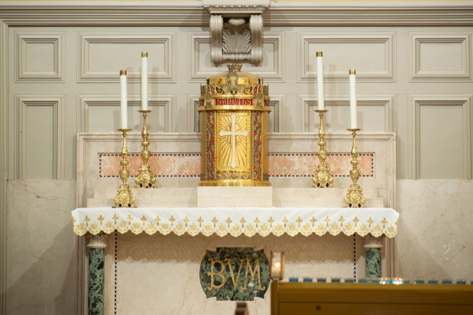 Ornate altar with a golden tabernacle, four tall candlesticks, and a decorative cloth inscribed with "BVM." Marble background.