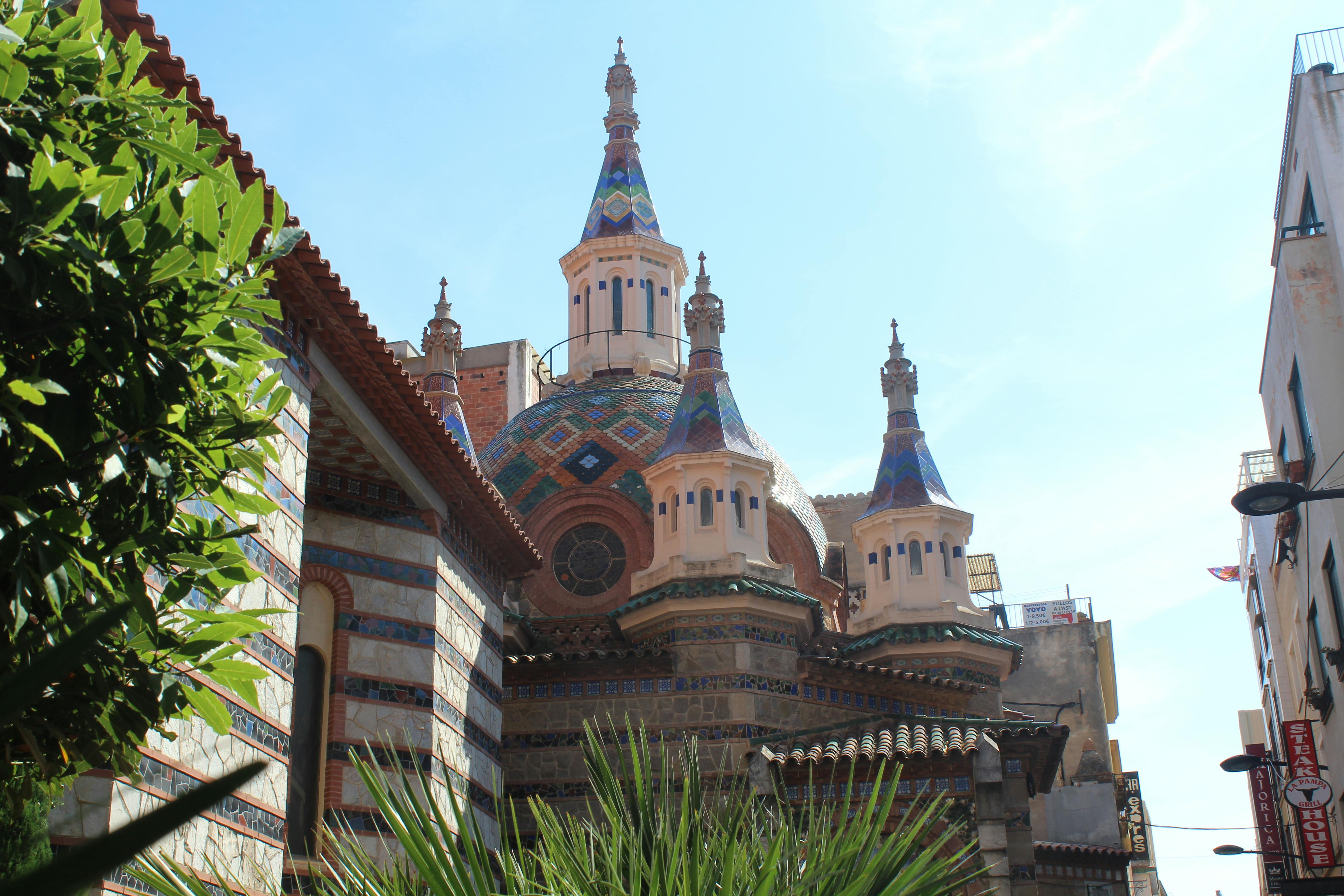 建筑采用彩色瓷砖穹顶和华丽的尖顶，周围环绕着晴朗的天空下的植物和部分可见的建筑物。
