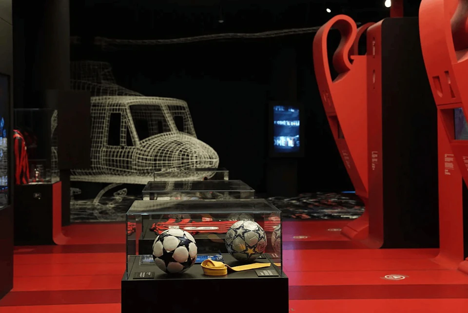 A display case containing two soccer balls and memorabilia, with a large red sculpture and a wireframe helicopter in the background.