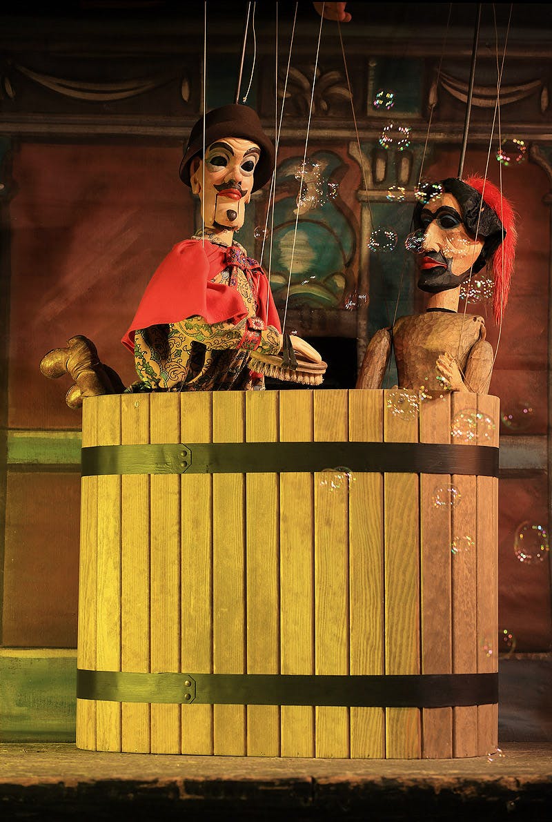 To marionetdukker inde i en trætønde med bobler, der flyder rundt, på en scene med en malet baggrund.
