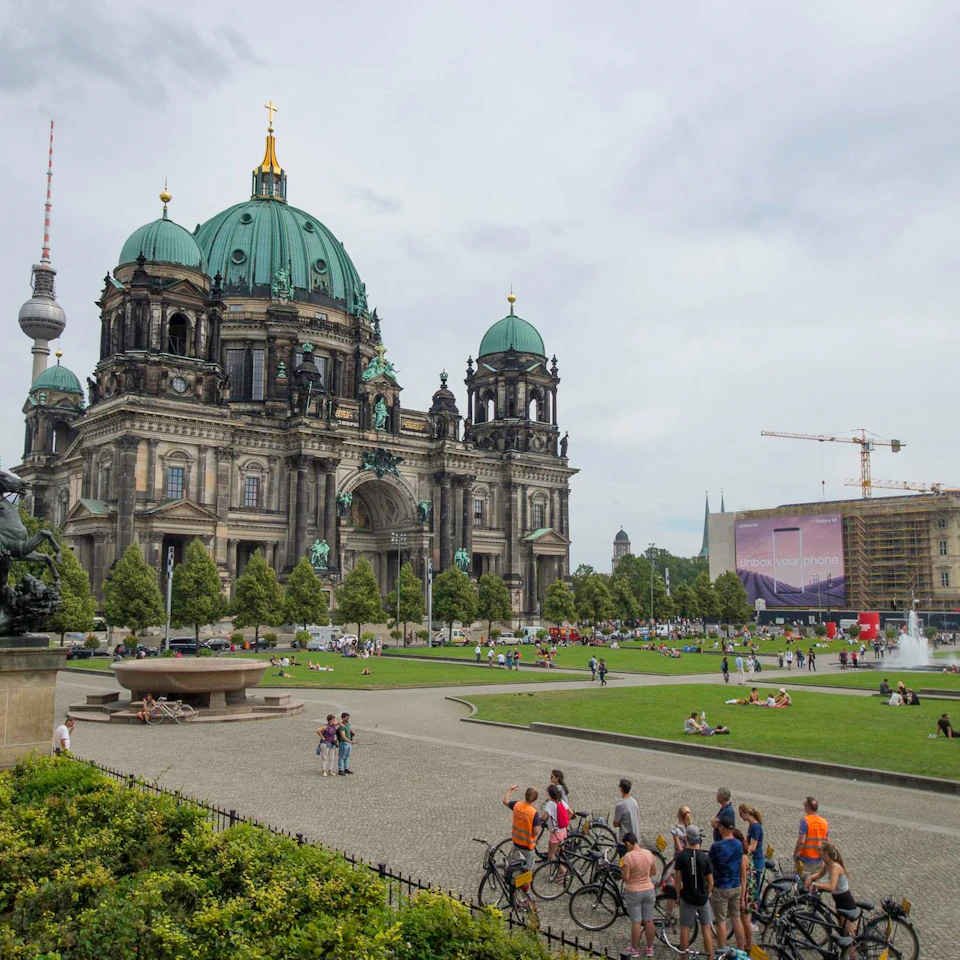 A large, ornate cathedral with green domes, a TV tower, and people walking or cycling in a park with a fountain nearby.