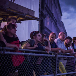 En grupp människor som lutar sig mot ett staket och tittar på något utanför skärmen, med en stadsbild och en molnig himmel i bakgrunden.