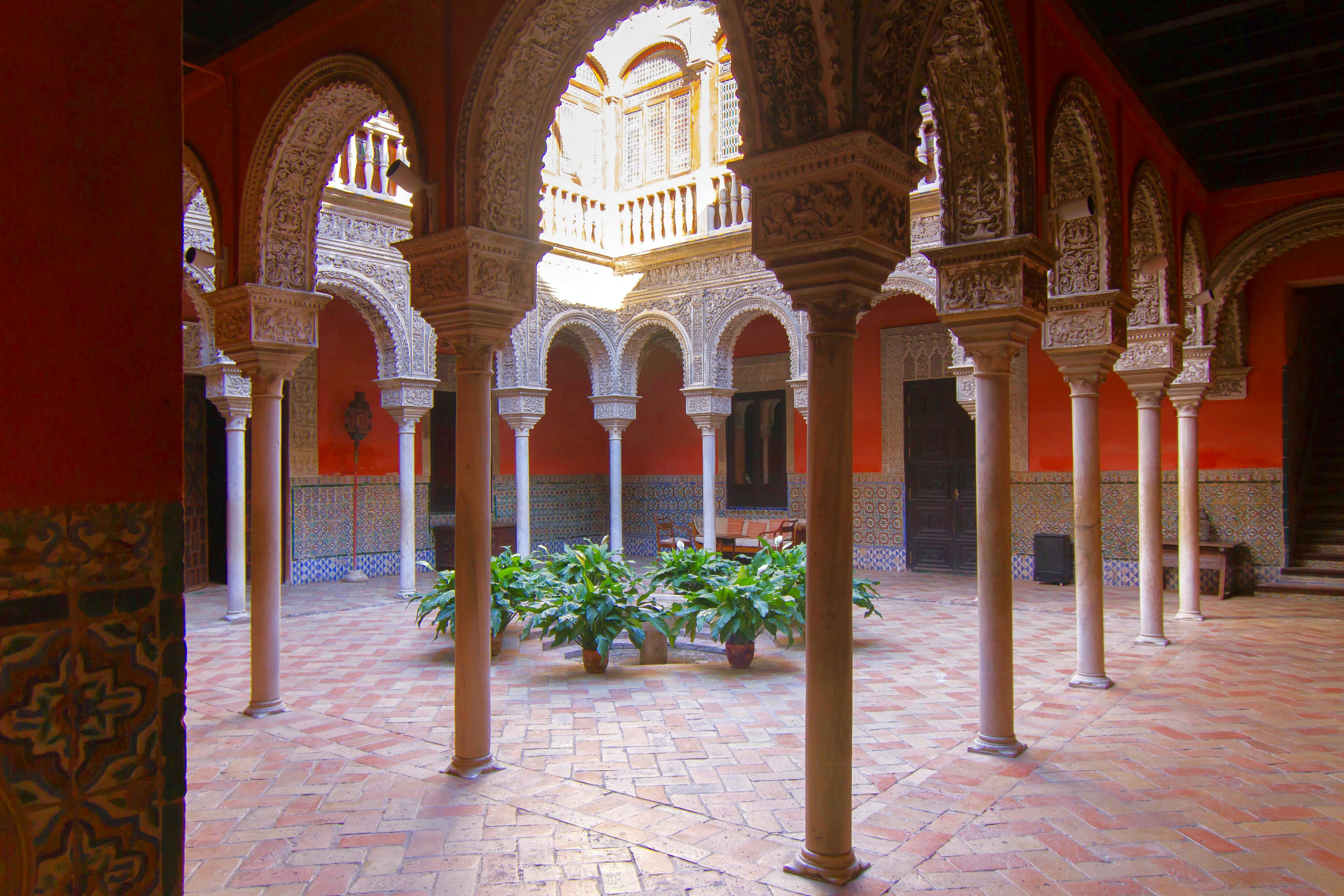 Ornate courtyard with intricately carved arches, tiled walls, potted plants, and a skylight, surrounded by columns on a brick floor.