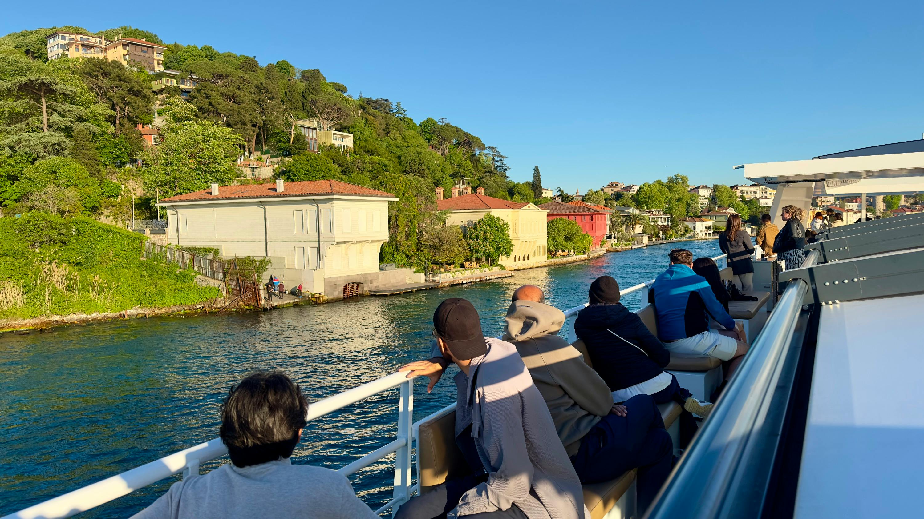 People seated on a ferry, enjoying the view of a canal lined with greenery and colorful houses under a clear blue sky.