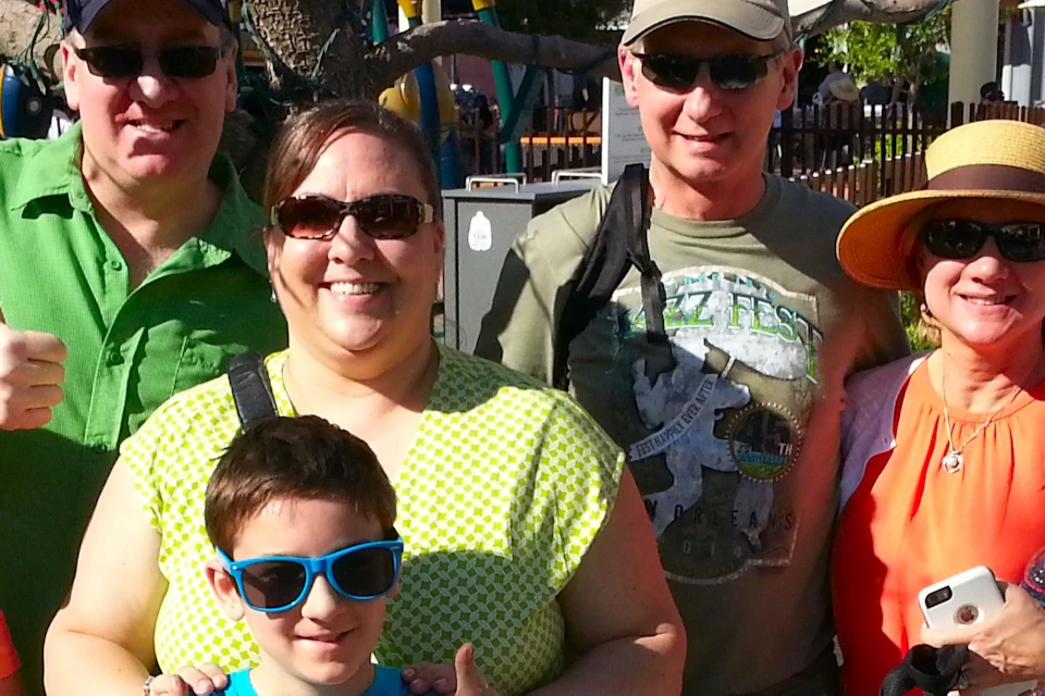 A group of seven people posing together outdoors on a sunny day, dressed in casual and colorful clothing.
