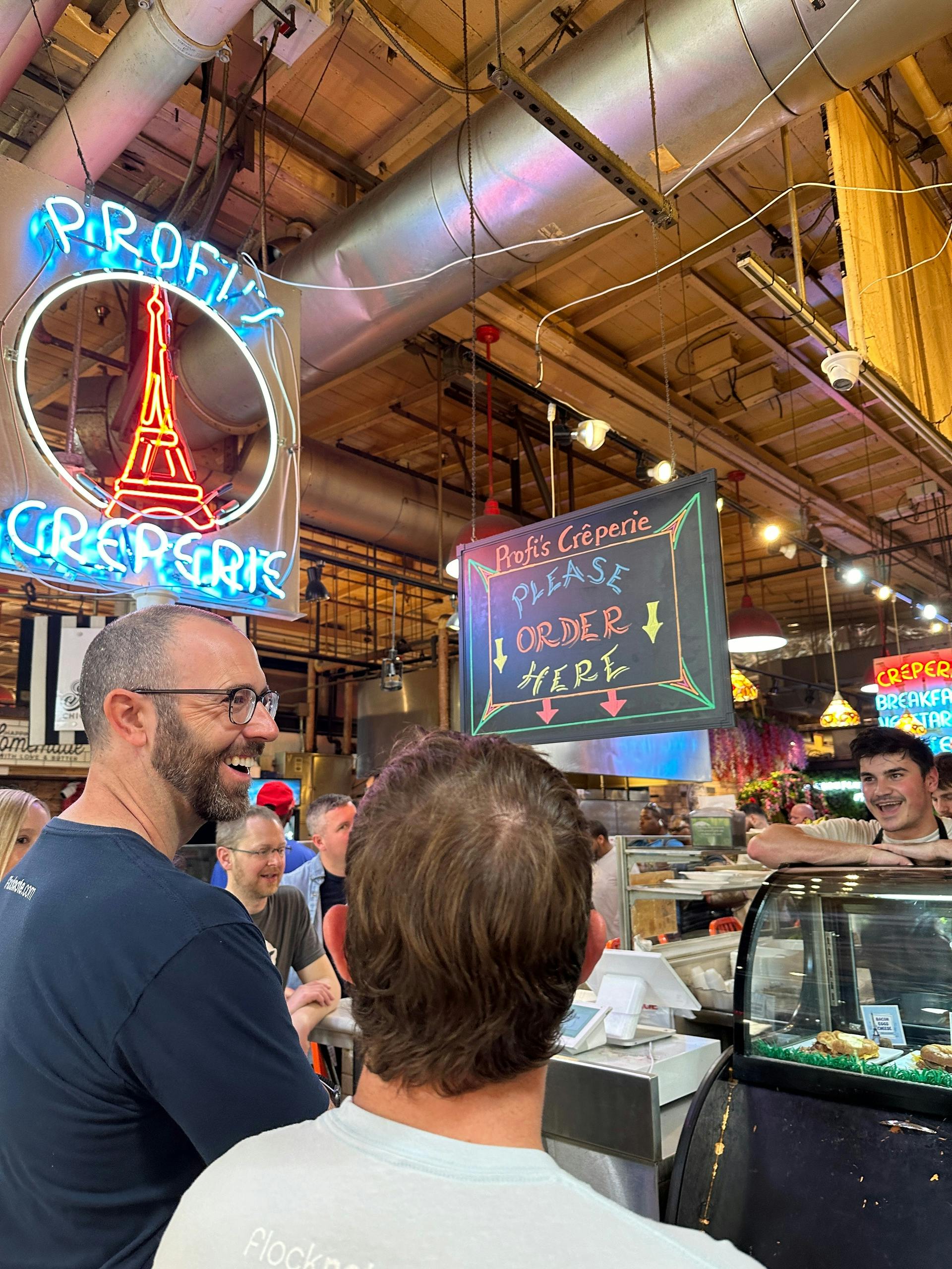 Menschen, die sich an einem Crêpe-Stand auf einem Markt mit Leuchtreklame und einer sichtbaren Menütafel über dem Kopf unterhalten.