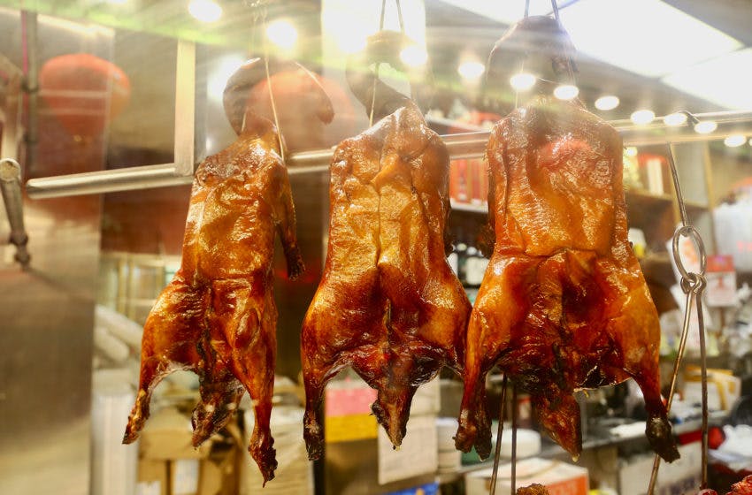 Drie hele geroosterde eenden hangend aan haken voor een verlichte etalage op een markt of in een restaurant.