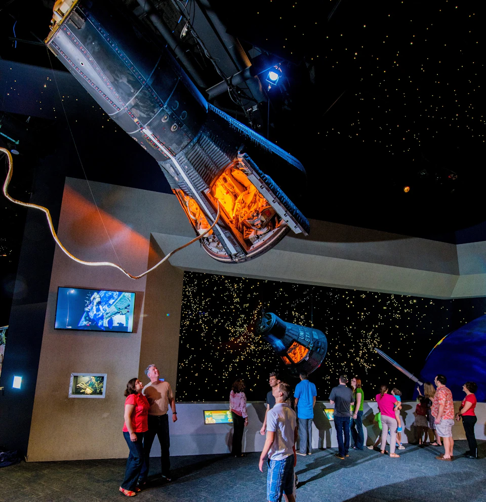 People viewing space exhibits, including suspended spacecraft, in a dimly lit museum with starry displays and informational screens.