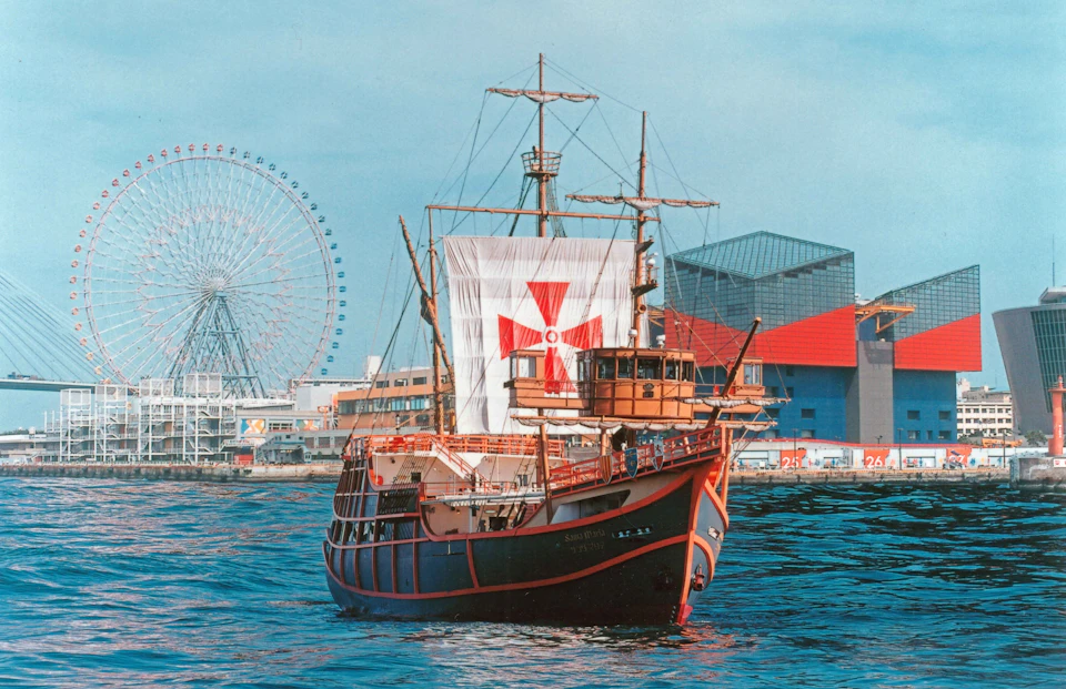 A sailing ship with a red cross on its sail is docked near a city waterfront, with a Ferris wheel and buildings in the background.
