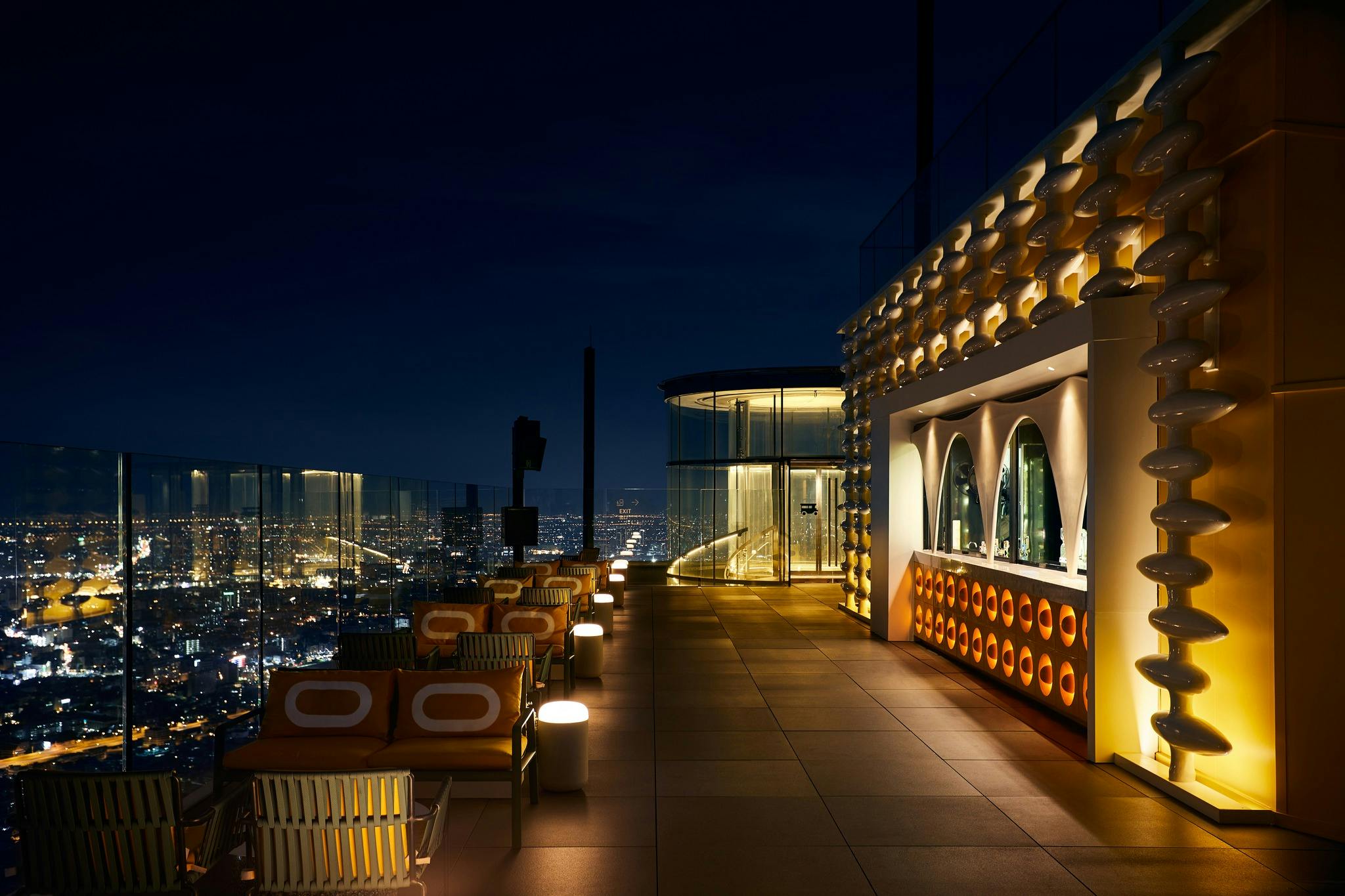 A rooftop lounge at night with city lights in the background, seating areas, and a modern bar with illuminated decor.