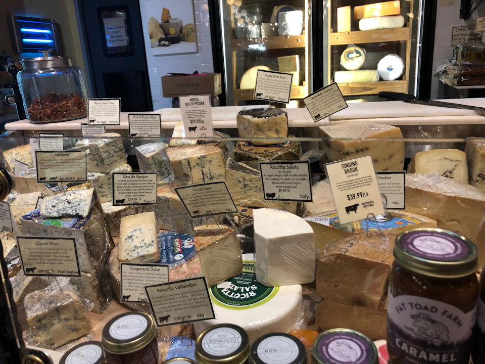A display of various cheeses with labels, including blue cheese and gouda, in a deli setting with jars of condiments in the foreground.
