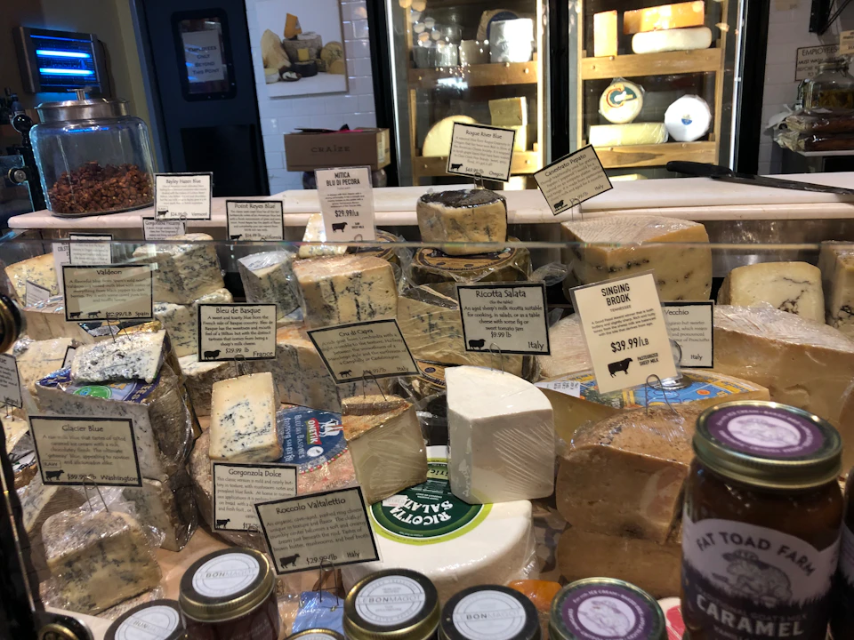 A display of various cheeses with labels, including blue cheese and gouda, in a deli setting with jars of condiments in the foreground.